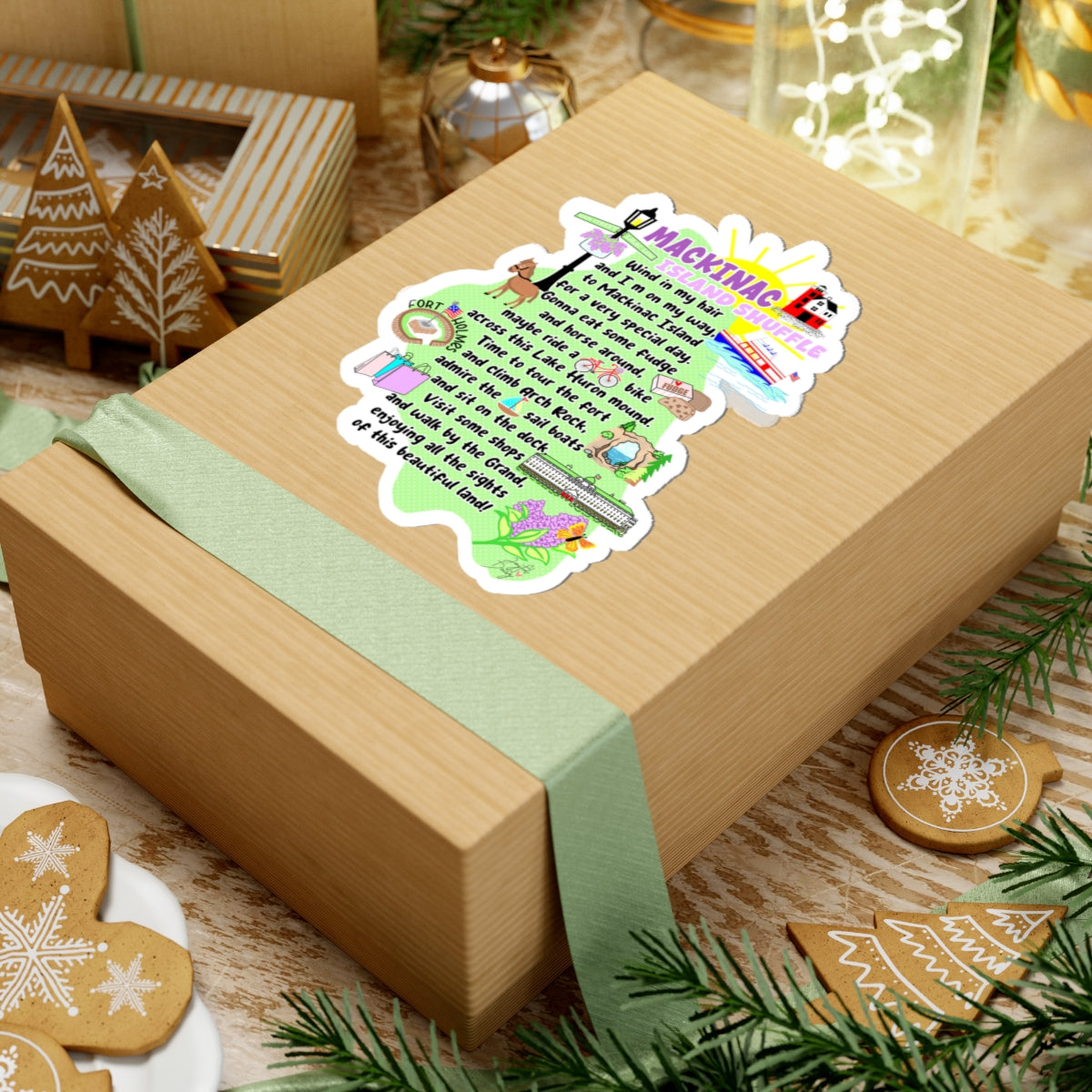 A kraft paper gift box tied with a pale green ribbon, decorated with a colorful Christmas-themed sticker that reads 'MACKINGNESS' and shows holiday illustrations. The box is styled on a rustic wooden surface with gingerbread cookies, pine branches, and other gift decorations.