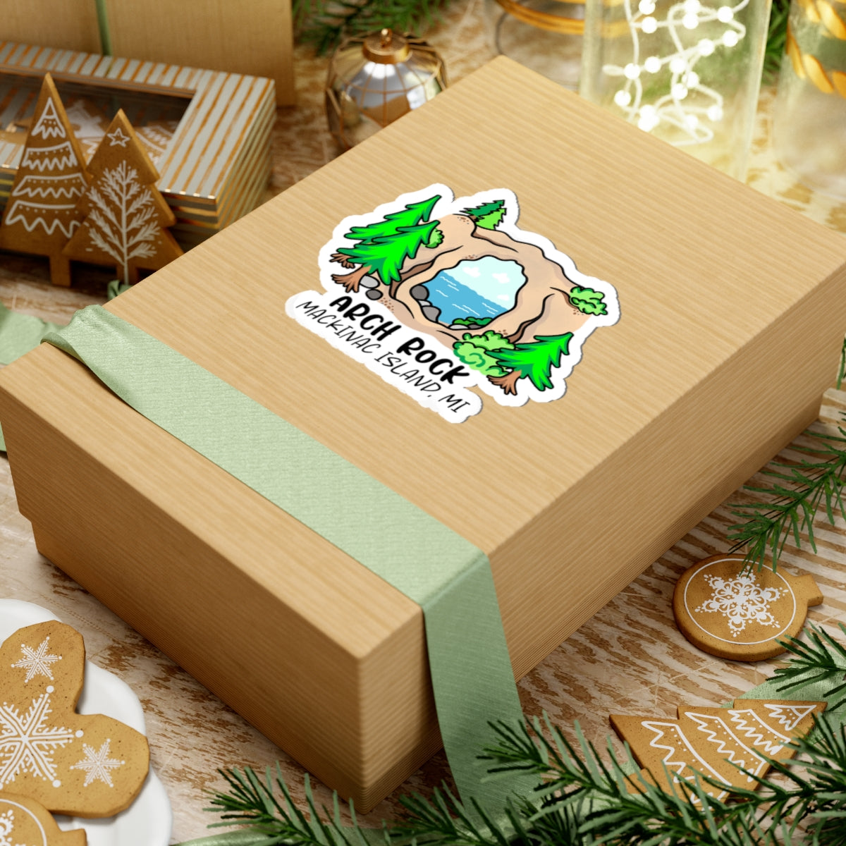 A square kraft paper gift box wrapped with a pale green ribbon, featuring a die-cut sticker that reads 'Apothrock Mountain Baking, ME' with a cartoon mountain and trees. The box is styled on a tabletop with holiday cookies, pine branches, and warm background decor.