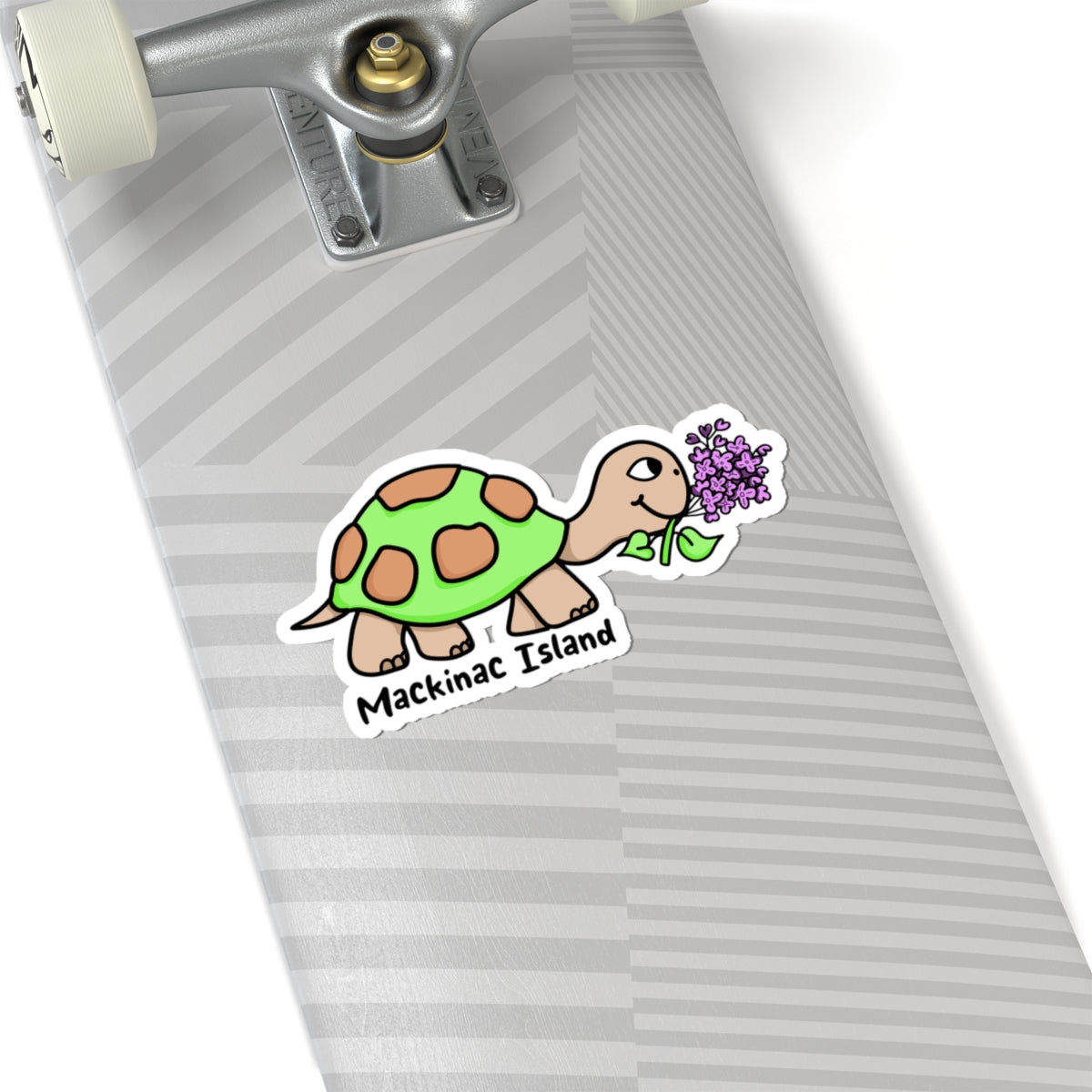 A die-cut sticker featuring a cartoon turtle eating purple flowers with the text 'Mackinac Island' below it, shown applied to the underside of a skateboard deck.