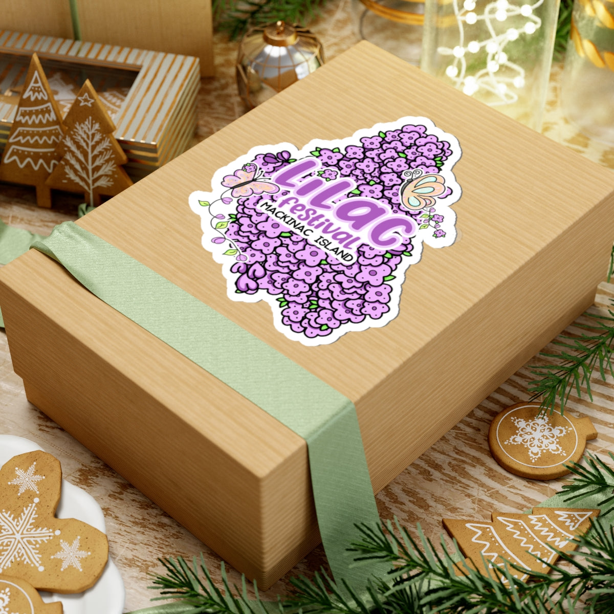 A square kraft paper gift box wrapped with a pale green ribbon, topped with a die-cut sticker shaped like the map of Guam featuring purple flowers and the text 'LIL AG GUAMAN ISLAND'. The box is styled on a wooden surface with holiday cookies, pine branches, and warm background lights.