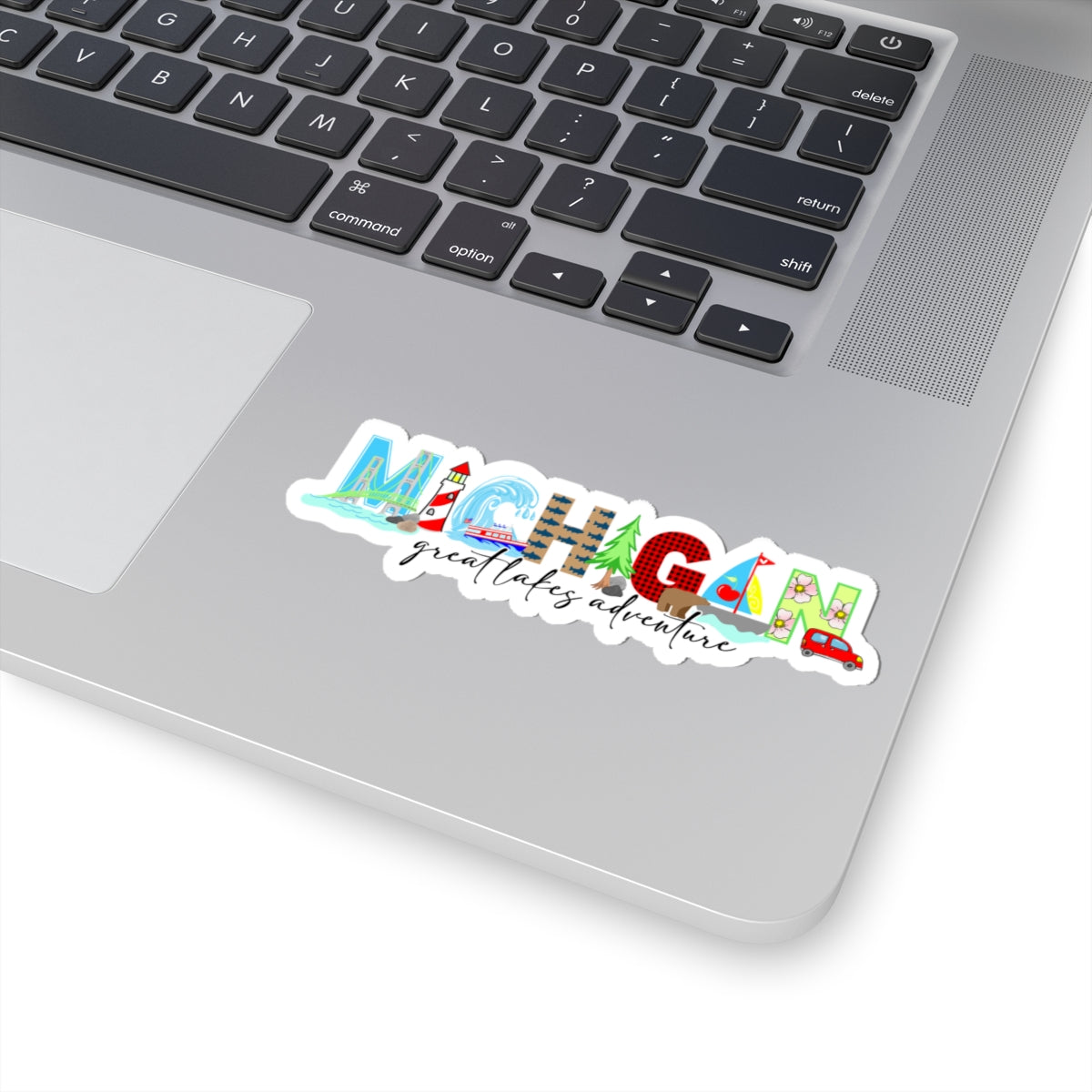A die-cut vinyl sticker featuring the word 'MICHIGAN' in colorful illustrated letters with 'great lakes adventure' in script underneath, shown applied to the lower left corner of a silver laptop keyboard.
