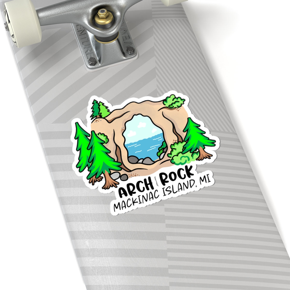 A die-cut sticker featuring a cartoon Arch Rock scene on a skateboard deck. The sticker shows green pine trees, a tan rock arch with blue water in the center, and the text 'ARCH ROCK MACKINAC ISLAND, MI'.