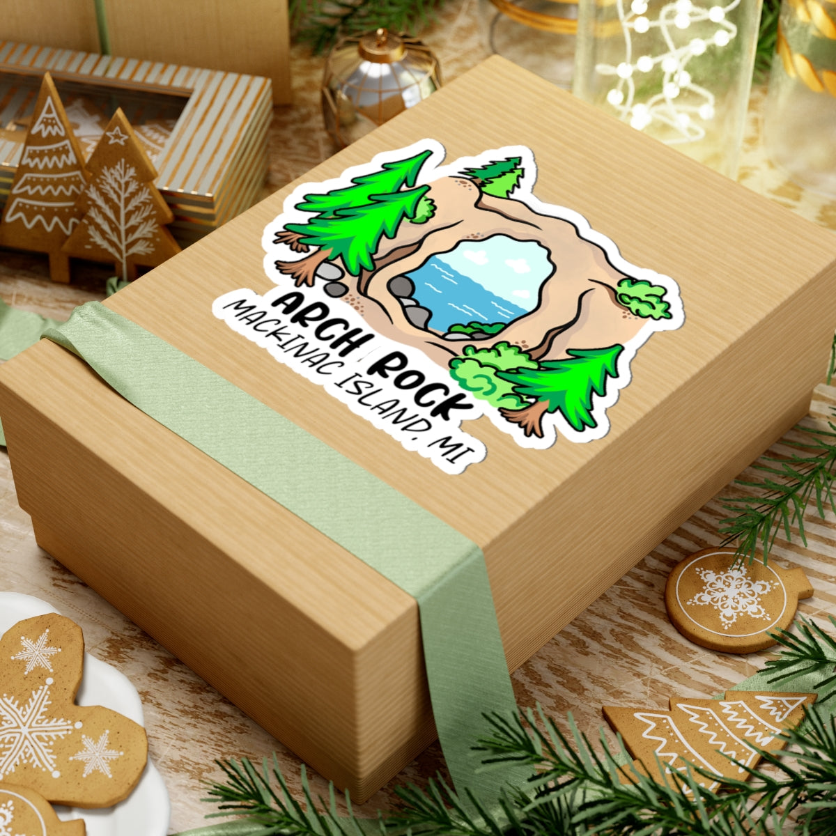 A kraft paper gift box tied with a pale green ribbon, featuring a die-cut sticker that reads 'ARCH ROCK MACKAWAC ISLAND MI' with a scenic illustration. The box is styled on a tabletop with holiday decorations, pine branches, gingerbread cookies, and warm background lights.