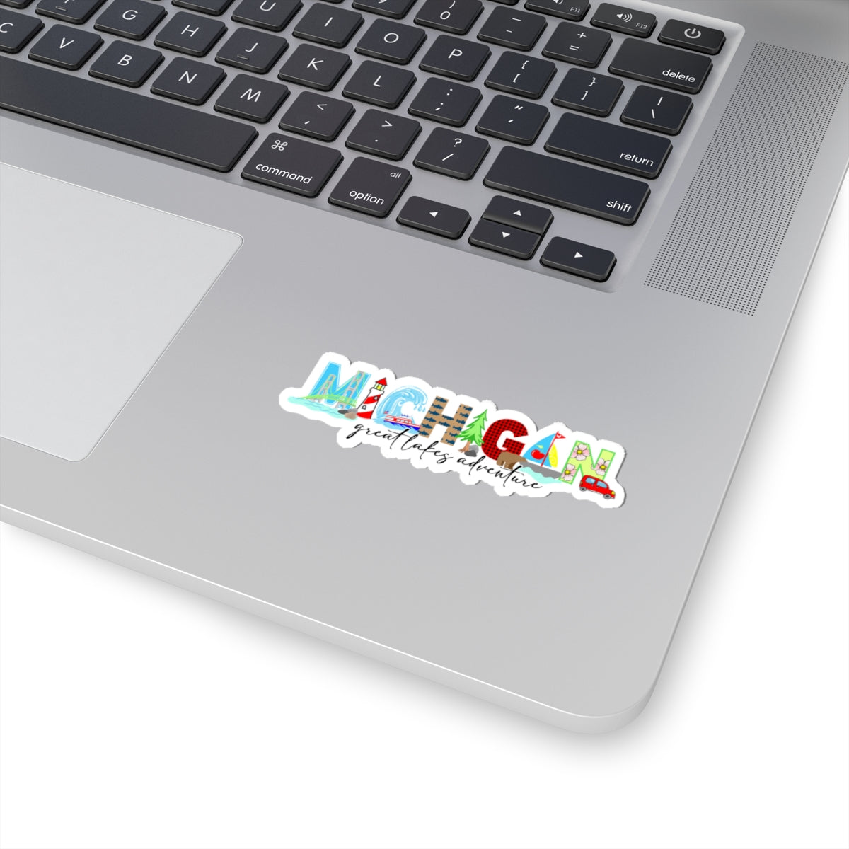 A colorful die-cut sticker reading 'MICHIGAN great lakes adventure' applied to the palm rest of a silver laptop. The sticker features a multicolor landscape design with blue, green, red, and yellow elements, shown on a clean white background.