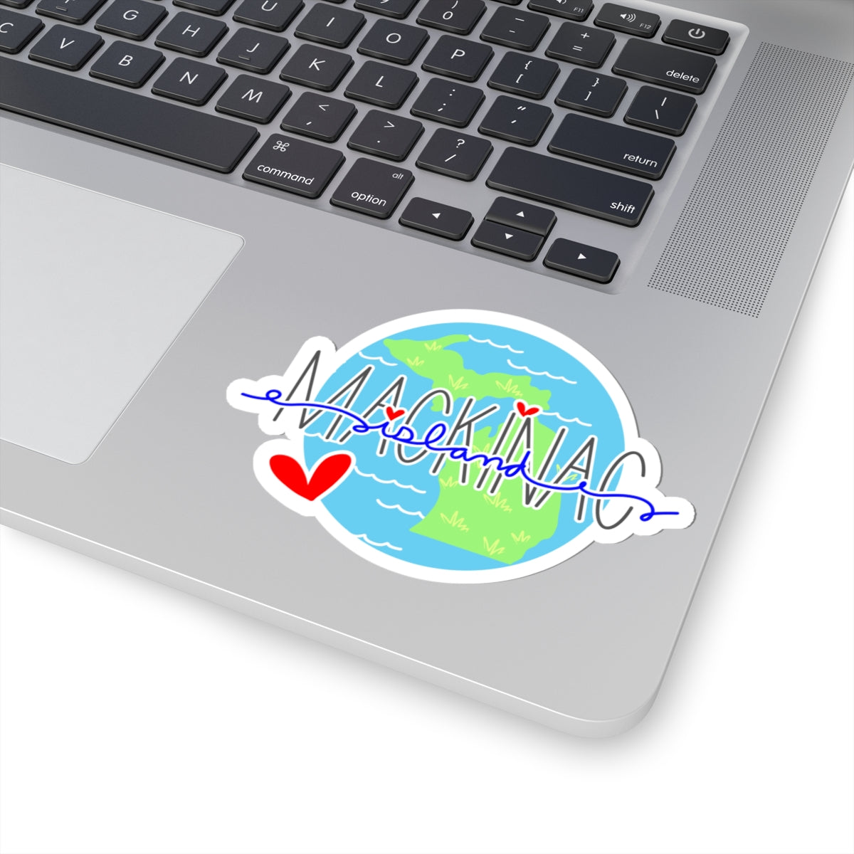 A die-cut vinyl sticker of the map of Michigan with 'MICHIGAN' written across it in blue script, decorated with red hearts, shown applied to the silver surface of a laptop.