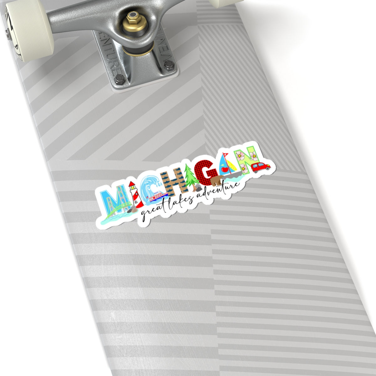 A die-cut sticker featuring the word 'MICHIGAN' in colorful illustrated letters and the phrase 'great lakes adventure' in black script, shown placed on the deck of a skateboard with a gray and white striped pattern.