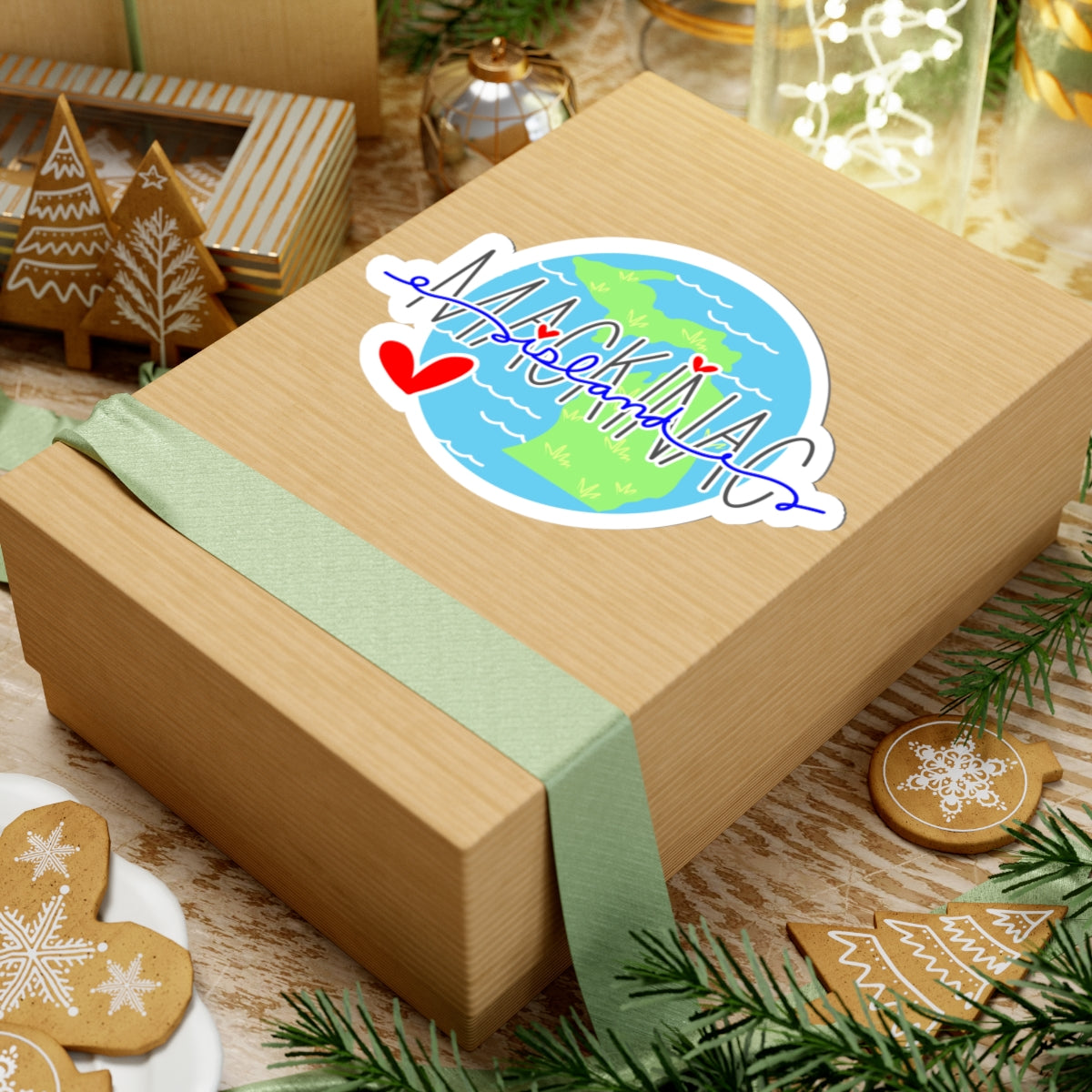 A square kraft paper gift box wrapped with a pale green ribbon and topped with a die-cut sticker showing a blue-and-green globe and the word "MACKENZIE" in blue script, surrounded by holiday decorations on a tabletop.