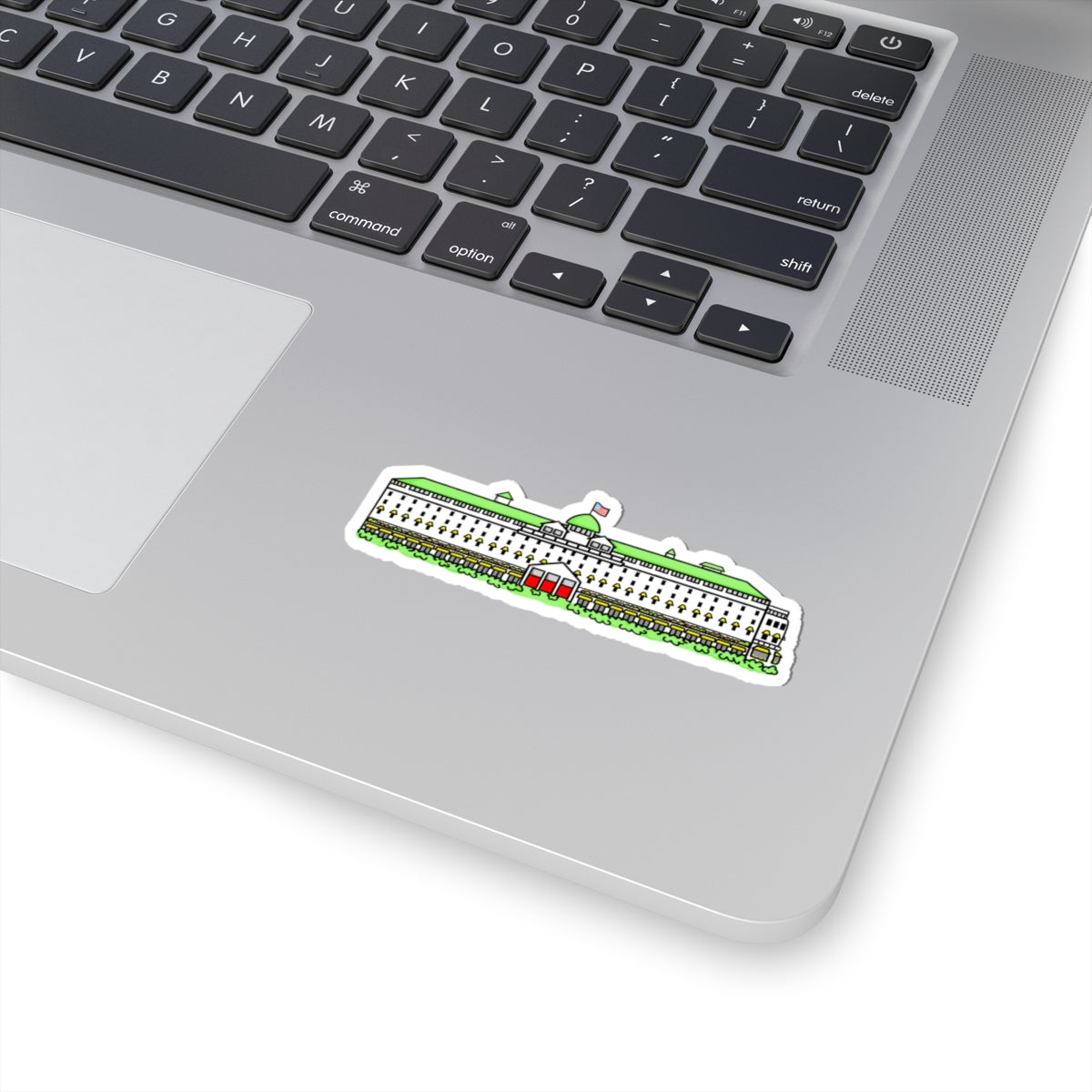 A die-cut sticker depicting the grand hotel on mackinac island michigan with a small red heart on its roof, shown applied to the silver palm rest of a laptop keyboard.