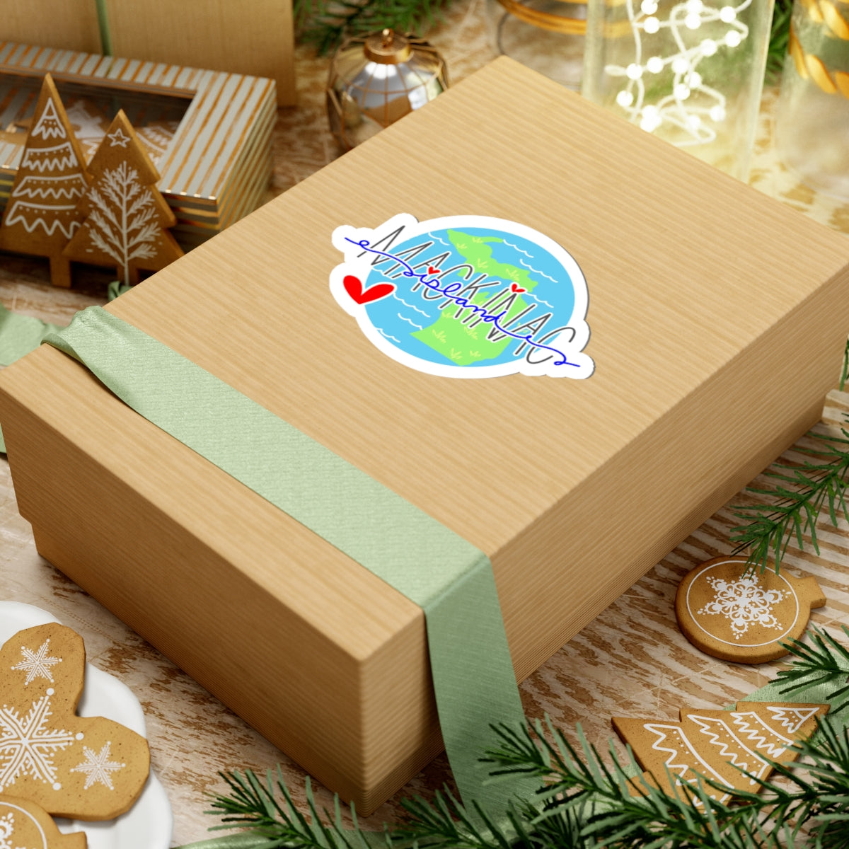 A kraft paper gift box wrapped with a pale green ribbon, decorated with a colorful 'Happy Holidays' globe sticker, set on a rustic wooden table surrounded by holiday decorations.