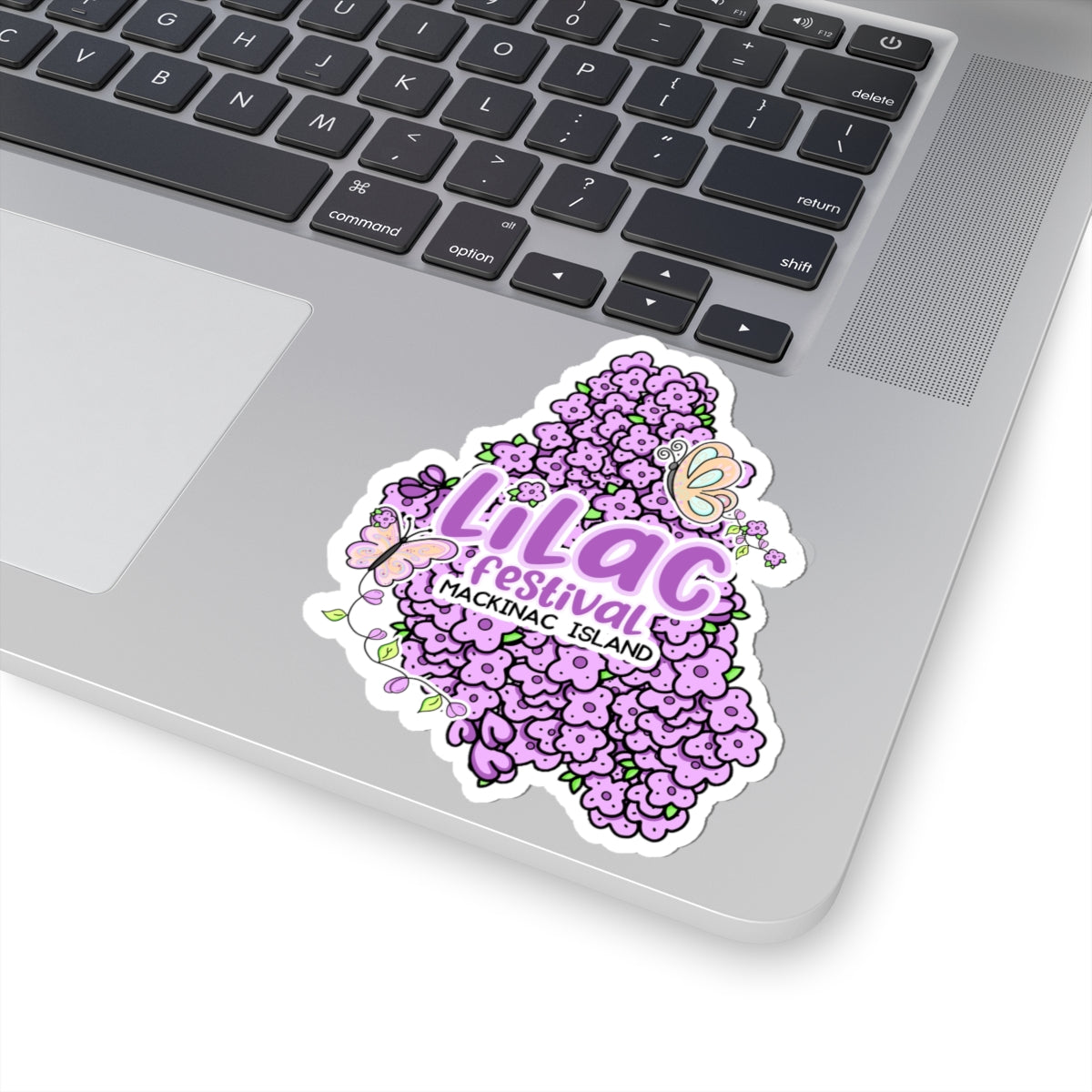A die-cut vinyl sticker of the Lilag Festival on a silver laptop trackpad. The sticker features a dense cluster of purple hydrangea flowers with a butterfly and the text 'Lilag Festival A HAKKINAG, ISLAND' in pink and purple lettering.
