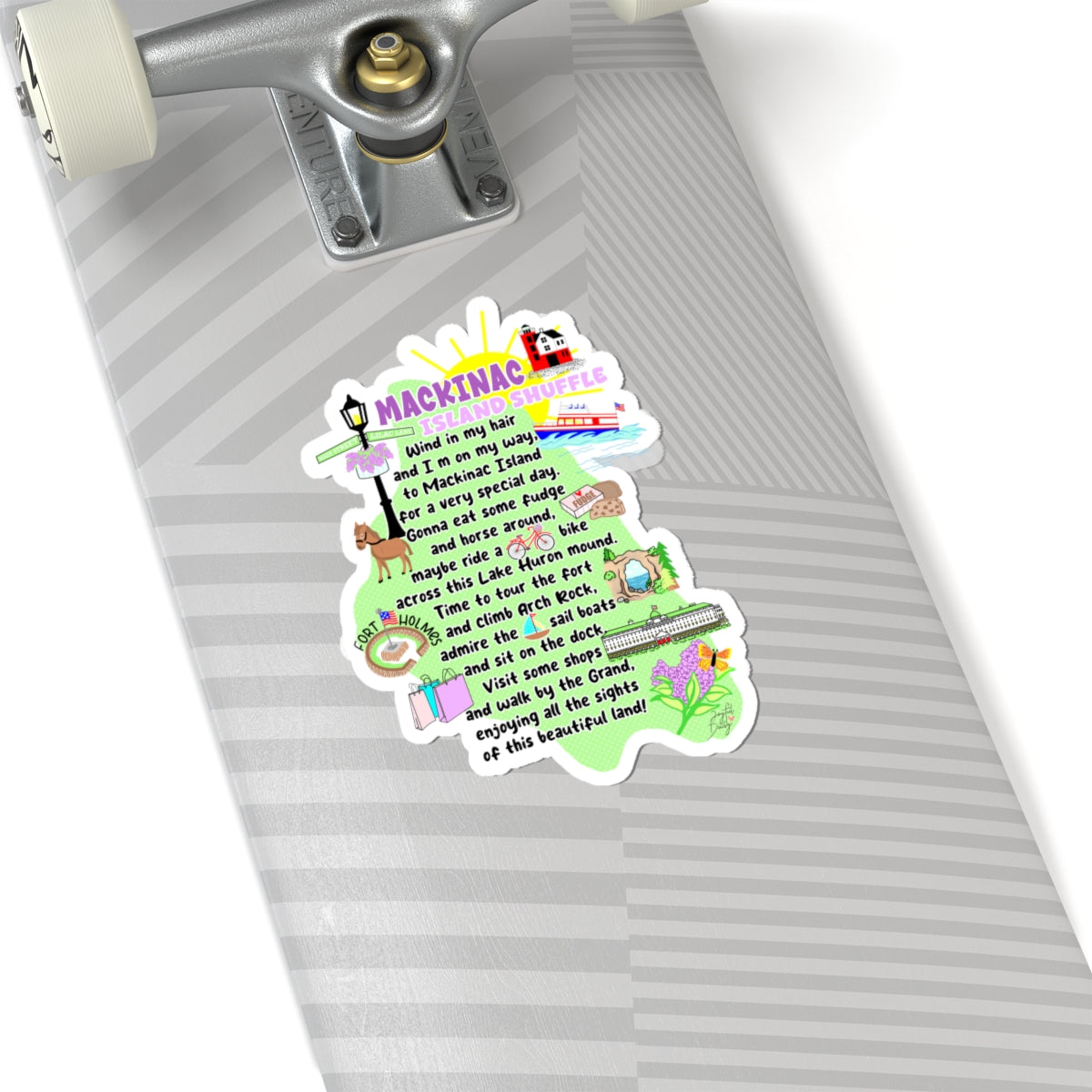 A die-cut sticker shaped like a skateboard deck, featuring a colorful illustrated design with the text 'MACKINAC IS' and a poem, shown applied to the underside of a silver skateboard.