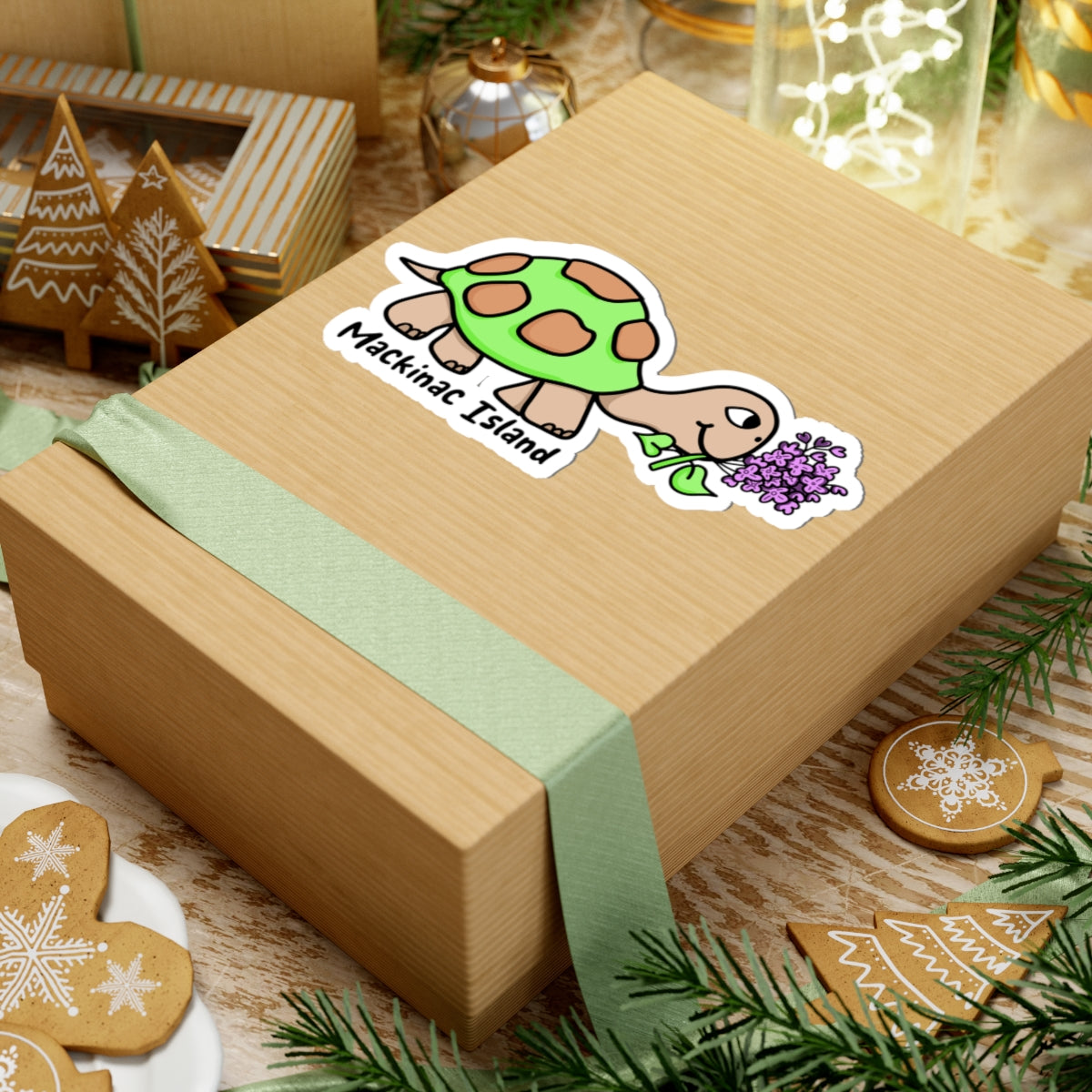 A square kraft paper gift box tied with a pale green ribbon, decorated with a cartoon turtle sticker labeled 'Makinac Island.' The box is styled on a tabletop with holiday cookies, pine branches, and warm decorative lights in the background.