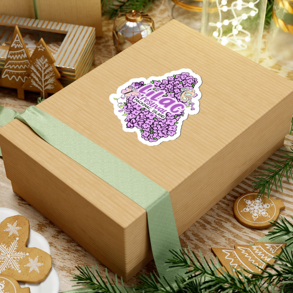 A kraft paper gift box wrapped with a pale green ribbon and topped with a die-cut sticker reading 'Hilag Michigan', styled on a tabletop with holiday cookies, pine branches, and warm decorative lights in the background.