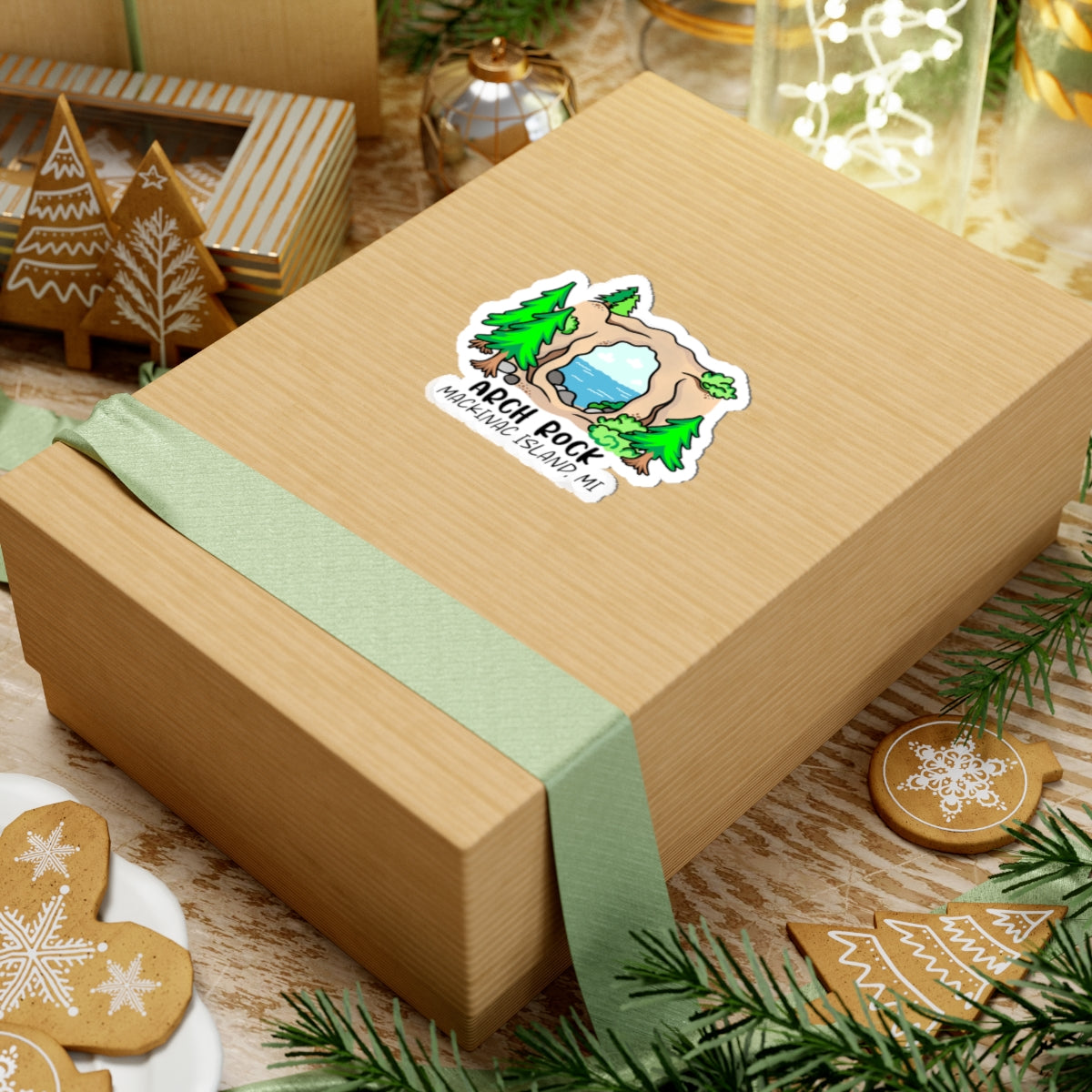 A square kraft paper gift box wrapped with a pale green ribbon, featuring a custom die-cut sticker that reads 'Aqua Pond' with a cartoon pond illustration. The box is styled on a rustic wooden table surrounded by holiday decorations, including gingerbread cookies, pine branches, and a small gingerbread tree ornament.