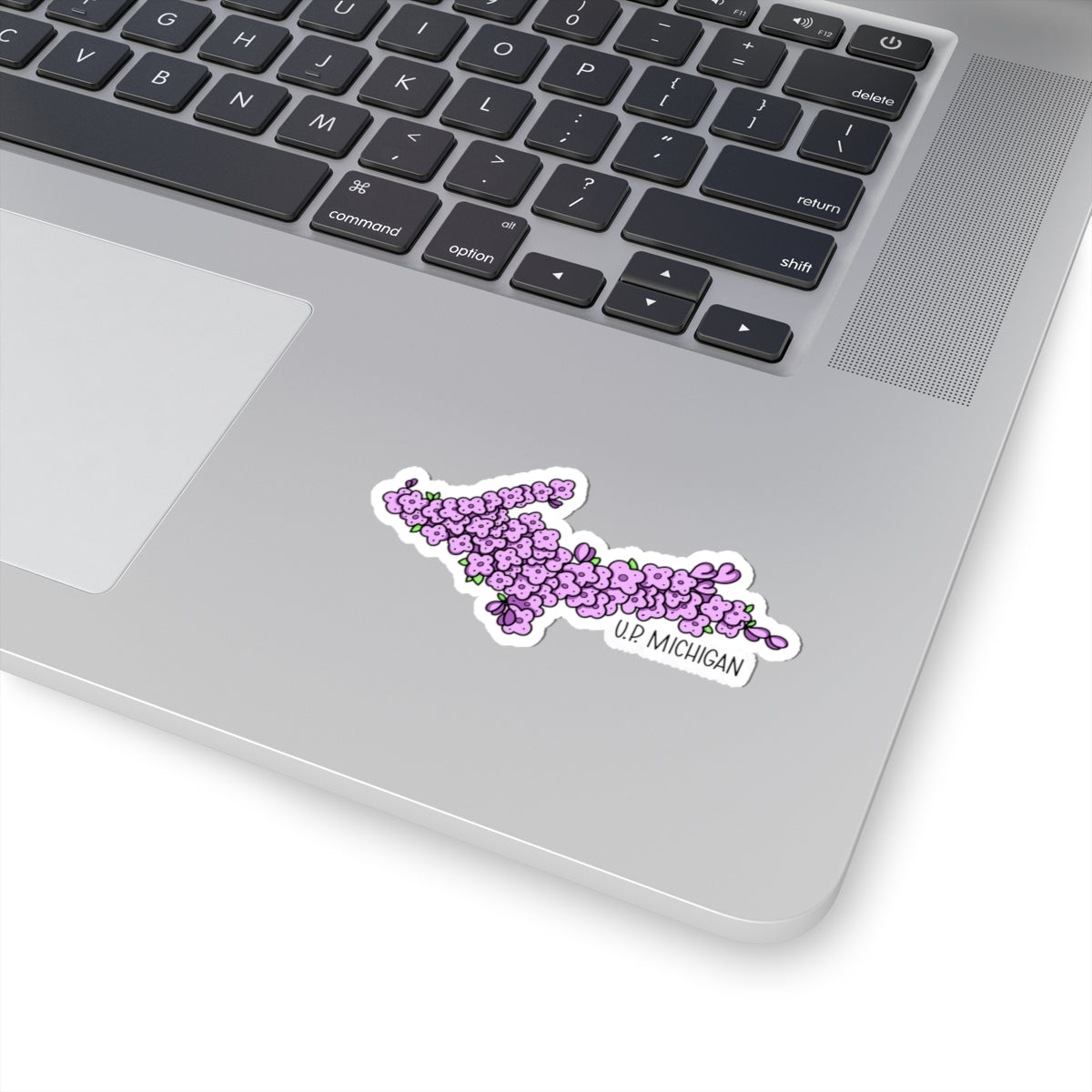 A die-cut vinyl sticker of the map of Upper Michigan, filled with purple lilac flowers and labeled 'UP MICHIGAN' in black text, shown applied to the palm rest of a silver laptop.