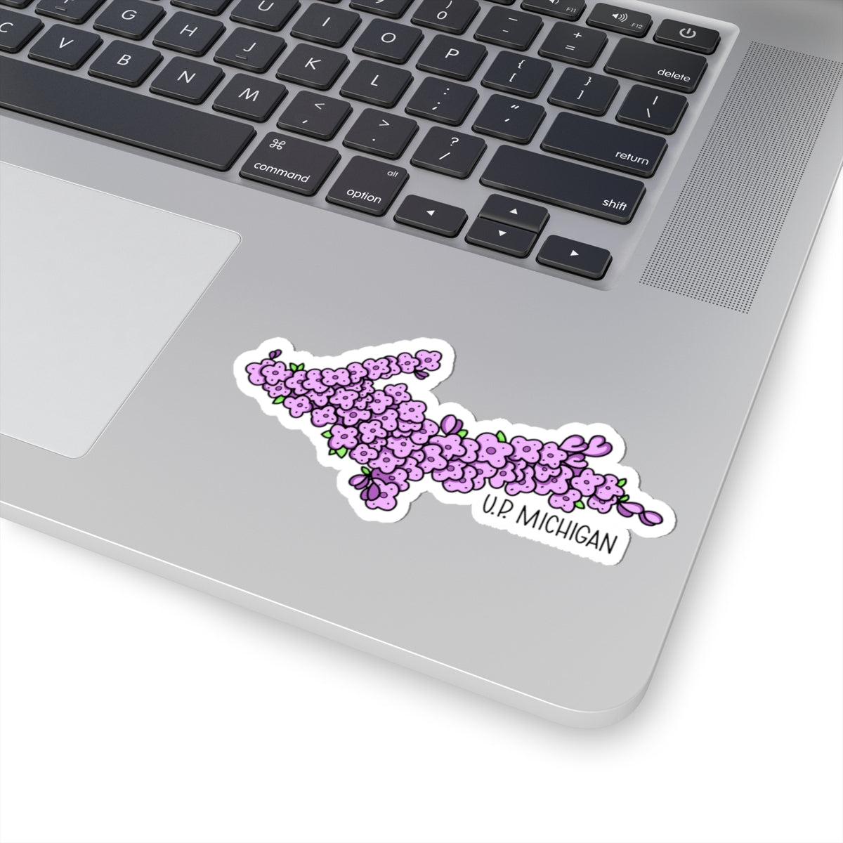 A die-cut vinyl sticker of the Upper Michigan map filled with purple wisteria flowers and the text 'UP MICHIGAN', shown applied to the silver palm rest of a laptop keyboard.