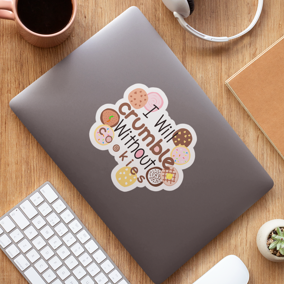 A die-cut sticker with the phrase 'I Will crumble without cookies' surrounded by illustrated donuts and cookies, shown applied to the back of a gray laptop. The laptop is photographed from above on a wooden desk alongside a keyboard, mouse, headphones, a mug, and a notebook.