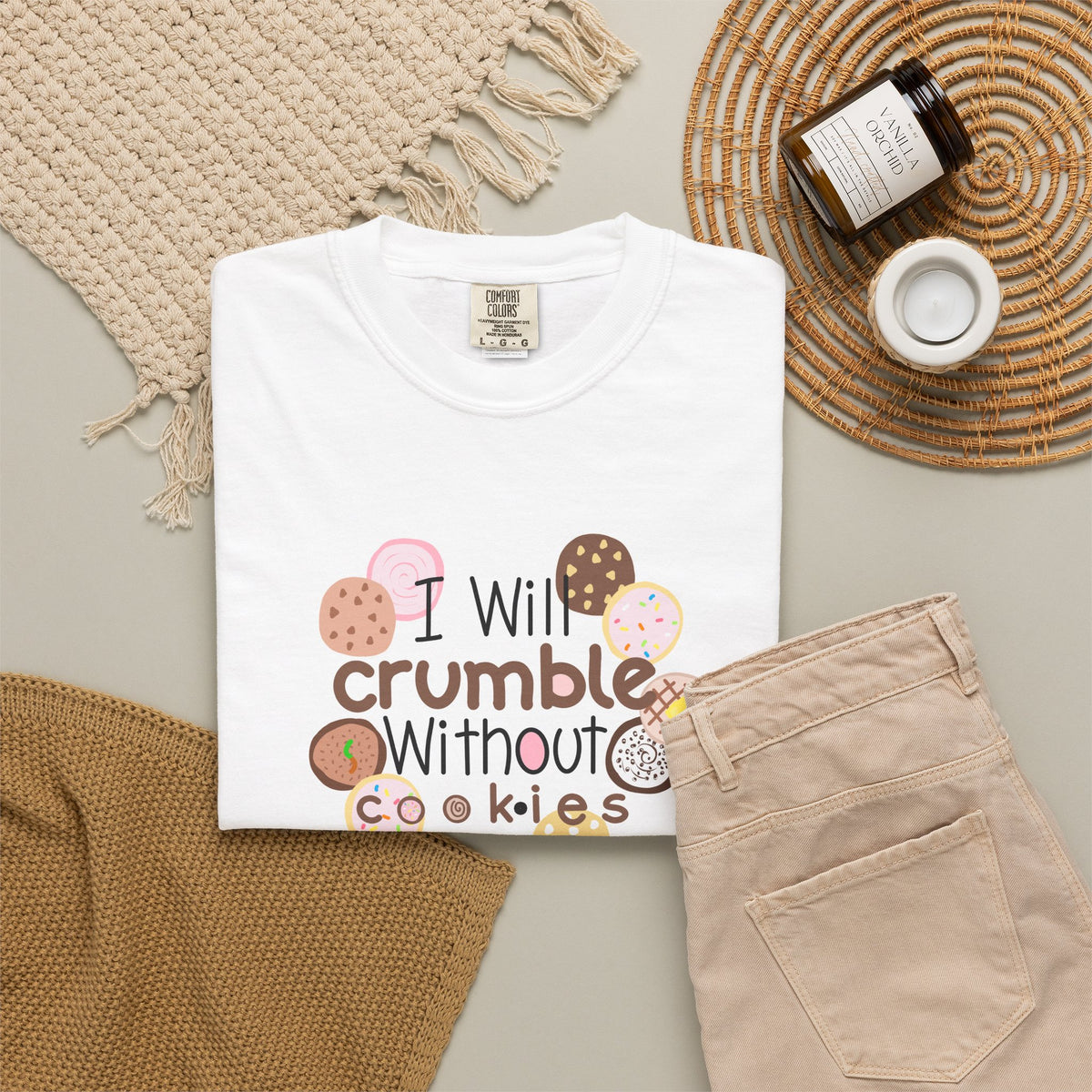 A folded white graphic T-shirt is arranged in a styled flat lay with beige pants, a woven blanket, a candle jar, and a small cup on a neutral surface. The shirt graphic reads 'I Will crumble Without cookies' with illustrated cookies in pink, brown, tan, and white tones.