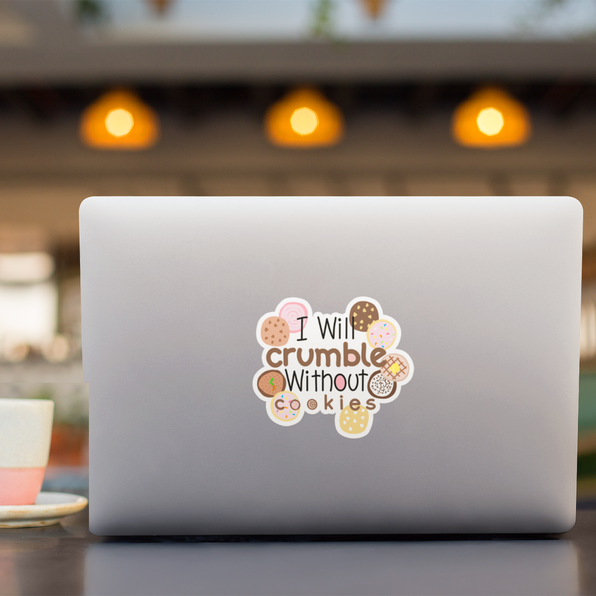A silver laptop on a table in a cafe setting, featuring a die-cut sticker on the back with the text 'I Will crumble Without cookies' surrounded by illustrated cookies.