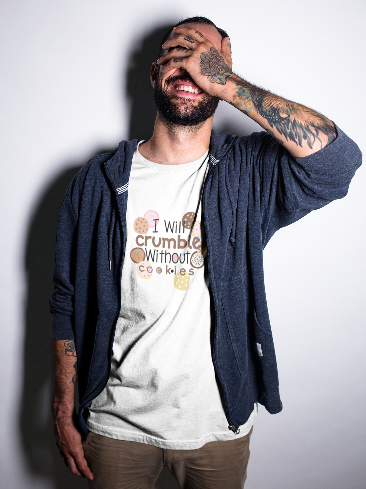 Crumble cookie tshirt on a man with tattoos and a beard covers his face with his hand, wearing a navy blue zip-up hoodie over a white graphic t-shirt that reads 'I Will crumble Without Cookies', and khaki pants, set against a plain light gray background.