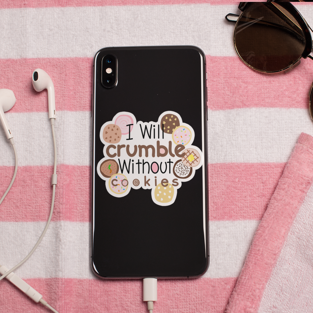 A black phone case with a cute cookie graphic and the text 'I Will crumble Without cookies' is displayed on a pink and white striped blanket. The scene is styled with white earbuds, sunglasses, and a charging cable.