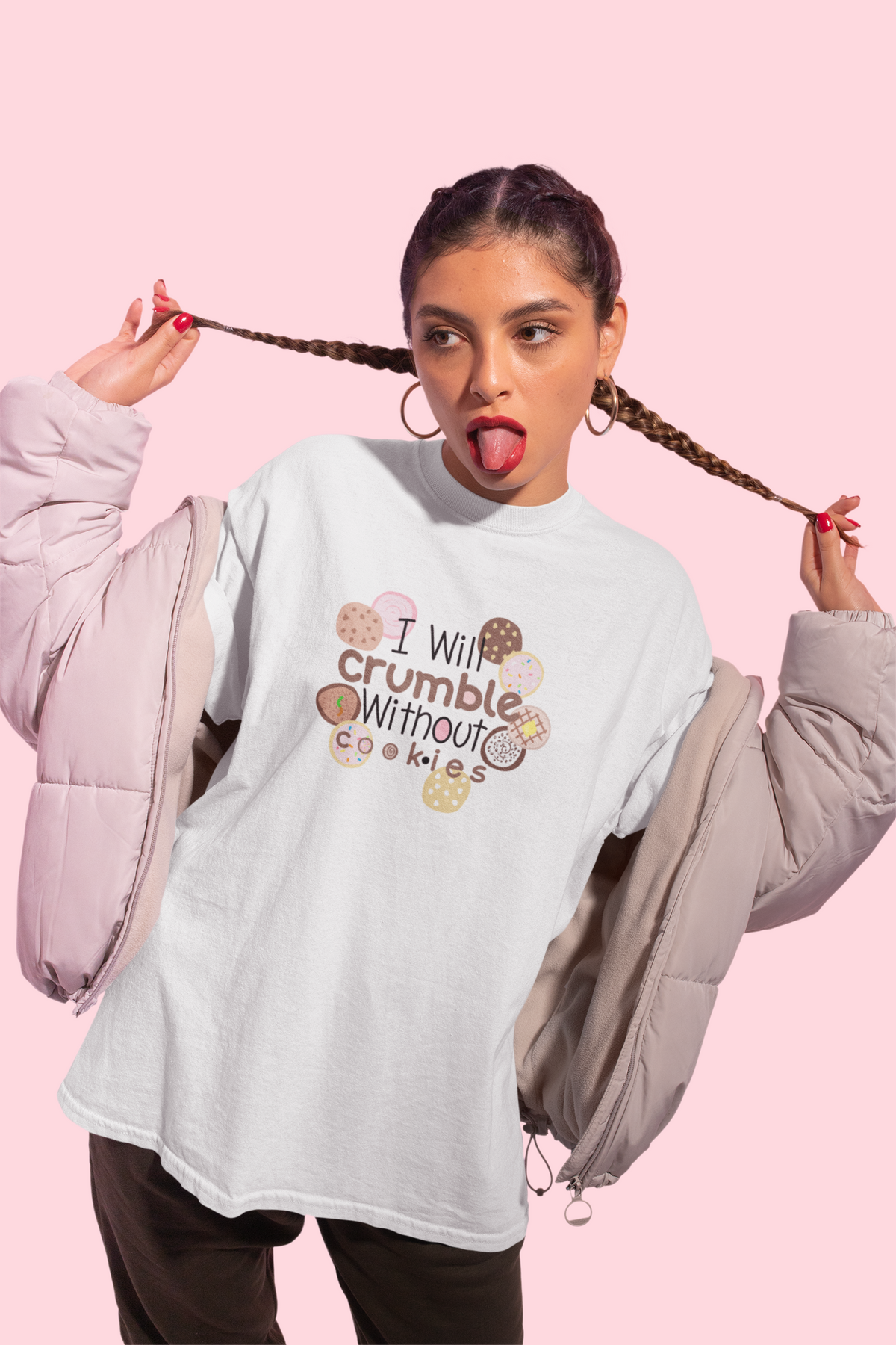 A woman modeling a light gray graphic t-shirt with the text 'I will crumble without cookies' surrounded by cookie illustrations. She is wearing an off-white puffer jacket draped off her shoulders and posing with her hair in two braids against a solid pink background.