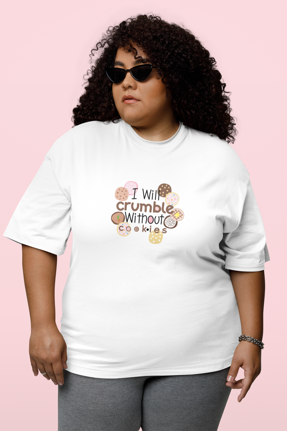 A woman with curly hair and sunglasses wearing a white graphic t-shirt that reads 'I Will crumble Without Cookies' surrounded by cookie illustrations, set against a solid pink background.