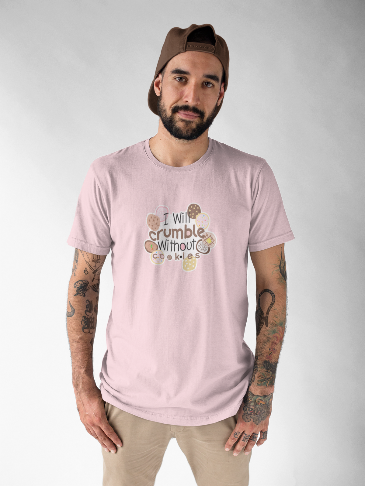 I Will Crumble for Cookies T-shirt, cute cookie lover gift for bakers A man wearing a light pink graphic t-shirt with the text 'I will crumble without a bit' surrounded by donut illustrations, styled with a brown baseball cap and khaki pants against a light gray background.